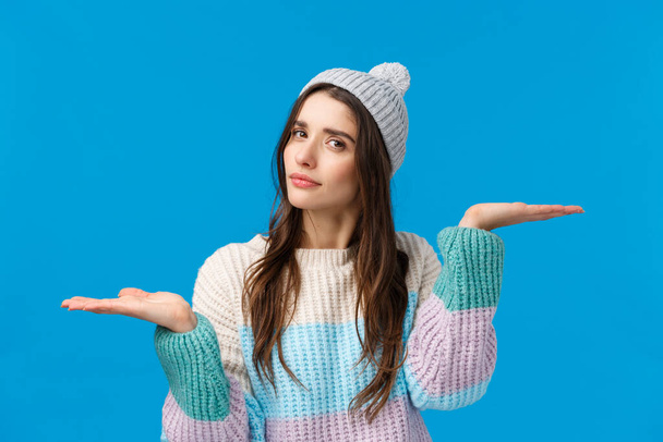 Girl troubled make choice, finally decided what buy, holding hands raised like weighing products, looking camera thoughtful, indecisive, asking help with picking winter holidays gift, blue background - Photo, Image