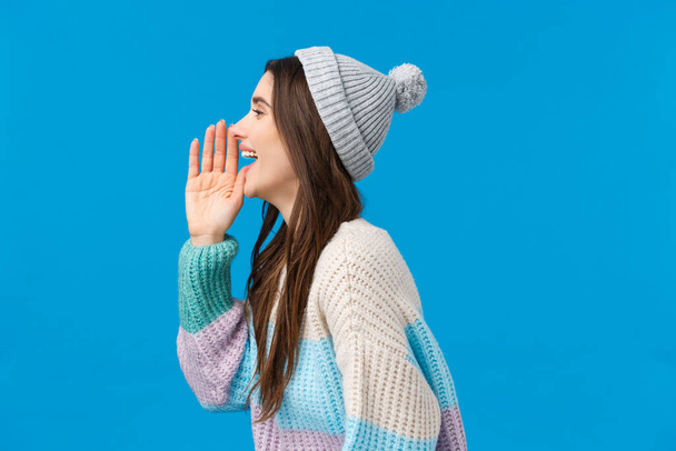 Are you coming or not. Happy and carefree outgoing smiling brunette girl in trendy winter sweater, hat, turn left and holding hand near opened mouth, calling someone, searching friend in crowd - Foto, imagen