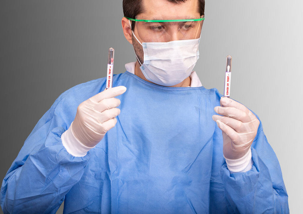 Mužský lékař v ochranných oděvech, rukavicích, masce a brýlích drží v rukou dvě zkumavky s negativním a pozitivním testem Covid 19 v laboratoři - Fotografie, Obrázek