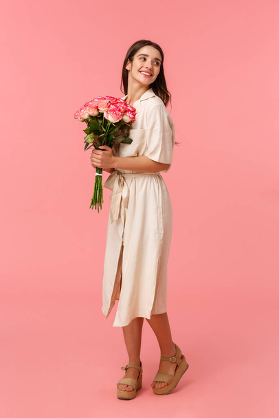 Full-length verticaal portret mooi brunette meisje op romantische datum ontvangen mooi boeket bloemen, holding rozen en staren achter met verrukt zacht en teder glimlach, staande roze achtergrond - Foto, afbeelding