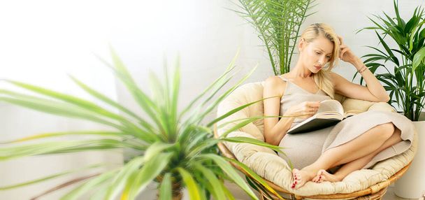 Woman relaxing, reading book in cozy room with green home plants. Boho home decor. - Фото, изображение