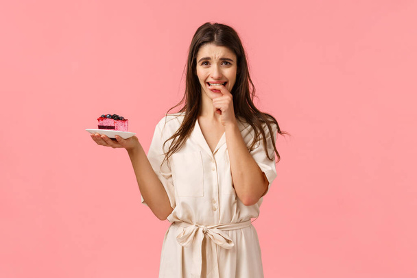 Maybe just one bite. Tempting and eager brunette woman want try tasty piece cake, holding dessert frowning and biting fingernails from desire to eat sweets, resist trying stick diet, pink background - Фото, зображення