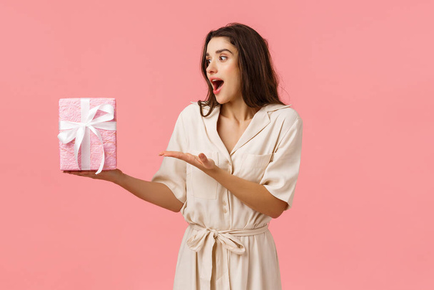 Girl likes surprises. Amused and happy cheerful young brunette woman in dress, receive gift box, pointing at present looking impressed and astonished as didnt expect such cuteness, pink background - Photo, Image