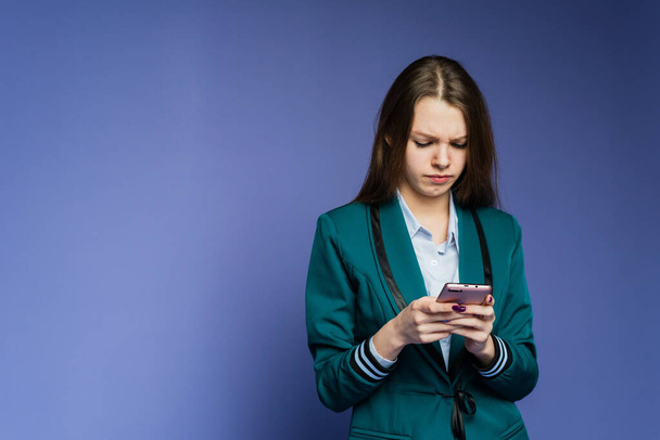 Een attent meisje met een mobiele telefoon staat op de blauwe achtergrond. Verwarrend meisje zoekt in telefoon op de achtergrond. Een meisje krijgt onaangename boodschap. - Foto, afbeelding