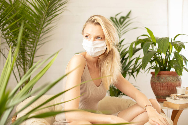 Blonde woman wearing medical face mask over green plants in home. - Foto, immagini
