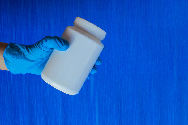 Ręka w niebieskie jałowe rękawice posiada biały plastikowy słoik na pigułki, koncepcja leczenia chorób, niebieskie tło. Ręka rękawiczki z lekiem - Zdjęcie, obraz