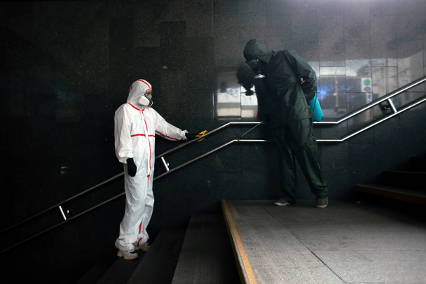 two people in protective suits Hazmat disinfect handrails from viruses and infections, disinfection of public places - Фото, изображение