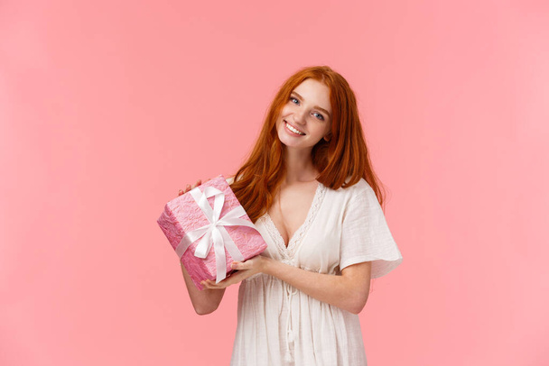 Lovely charismatic european woman with red curly hair, tilt head and smiling, congratulating with birthday, celebrating, attend b-day party giving her gift, holding present, pink background - Foto, Bild