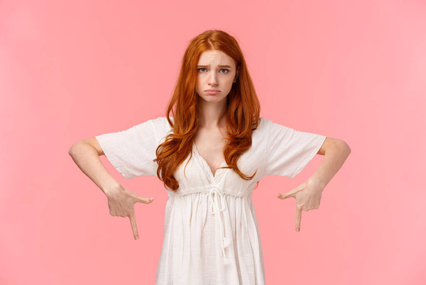 Несчастная, грустная рыжая девушка в белом платье чувствует себя неловко, указывая на что-то разочаровывающее, показывая нижнюю рекламу, указывая пальцами вниз и дуясь, розовый фон
 - Фото, изображение