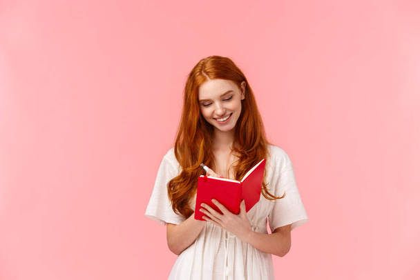 Schöne, sinnliche und stylische rothaarige Mädchen, die Listen erstellen, lernen, Hausaufgaben schreiben oder Termine in einem niedlichen roten Notizbuch schreiben. lächelnd, verträumt auf Noten schauend, stehend, rosa Hintergrund erfreut - Foto, Bild
