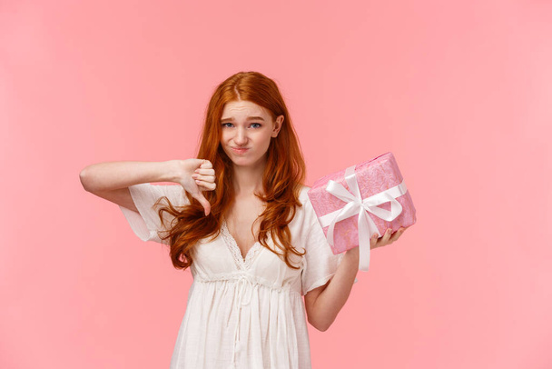 Disappointment, celebration and people concept. Waist-up portrait cute and picky redhead girl feeling awkward as holding cute box with gift, make thumb-down gesture and cringe, dislike present - Фото, зображення