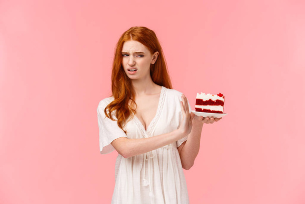 Take it away, gross. Ignorant and picky spoiled redhead birthday girl refusing eating dessert, holding plate with cake and making stop, rejection gesture, turn away with disgust, pink background - Photo, Image