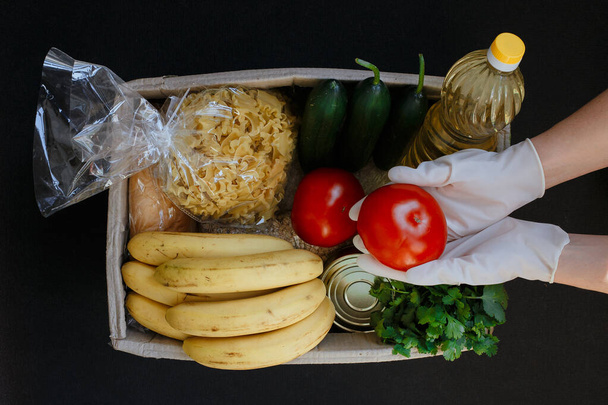Ręka w gumowej rękawiczce trzyma pomidora. Pudełko z jedzeniem. Owoce, warzywa, konserwy, makaron i olej słonecznikowy w pudełku. Pomoc społeczna w zakresie żywności. - Zdjęcie, obraz