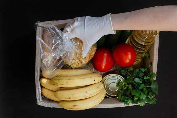 Ένα χέρι σε ένα λαστιχένιο γάντι κρατάει ντομάτα. Ένα κουτί δωρεών με φαγητό. Φρούτα, λαχανικά, κονσερβοποιημένα τρόφιμα, ζυμαρικά και ηλιέλαιο σε κουτί. Κοινωνική βοήθεια με τα τρόφιμα. - Φωτογραφία, εικόνα