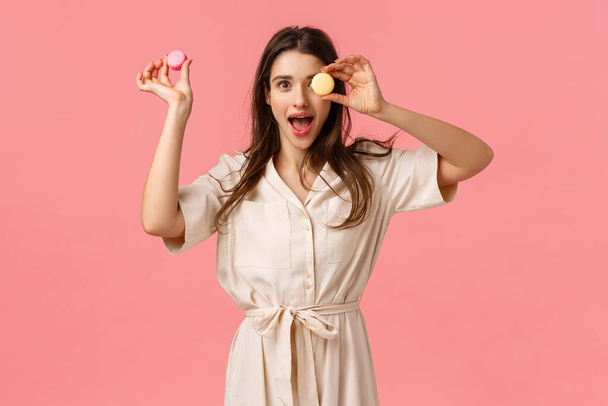 Excitement, holidays and sweet desserts concept. Elegant attractive young woman enjoy eating delicious food, holding macarons on on eye, open mouth gasping amused, standing pink background - Foto, Imagen