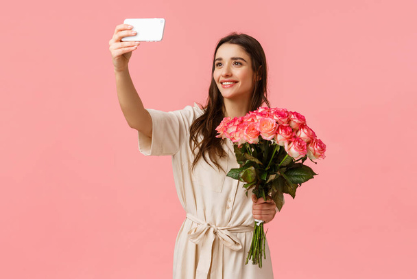 誕生日、休日、ロマンスの概念。魅力的な若い白人女性が配達を受け、ギフトの花を手に入れ、美しい花束でスマートフォンで自撮り、幸せなピンクの背景を立って - 写真・画像