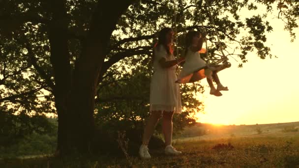 Mama schaukelt gesunde kleine Tochter auf Schaukel unter Baum in der Sonne. Eine Mutter spielt mit ihrem Kind, sie schwingen an einem Seil auf einem Eichenzweig im Wald. Die freie Familie hat Spaß im Park. glückliche Kindheit, Familie - Filmmaterial, Video