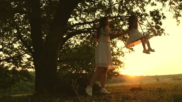 Moeder rockt gezond dochtertje op schommel onder boom in de zon. een moeder is aan het spelen met een kind ze zwaaien aan een touw op een eik tak in het bos. gratis familie heeft plezier in Park. gelukkige jeugd, familie - Foto, afbeelding