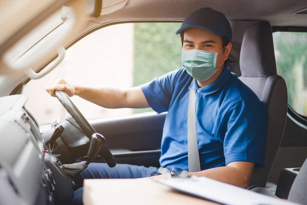 Young asian delivery man wearing protective face mask driving his van with packages on the front seat due to Coronavirus disease or COVID-19 outbreak situation in all of landmass in the world. - 写真・画像