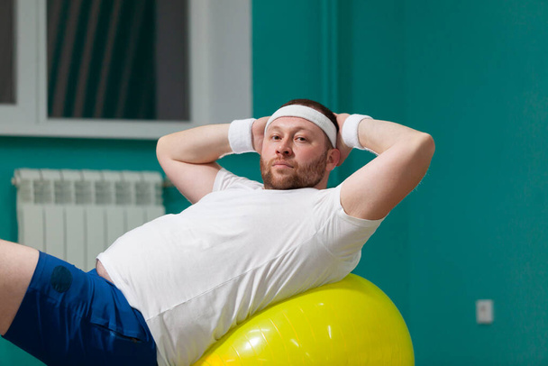 Overweight man is lying on a fitness ball during group fitness classes. Fat man looks disappointed because of bad result of weight loss trainings - Photo, image