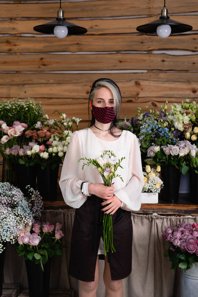 Business suffers during quarantine. Woman businessman engaged in the sale of color. One stands in an empty store holding flowers in her hands. A face shield is worn. Concept of small business losses during quarantine. - Foto, Imagen
