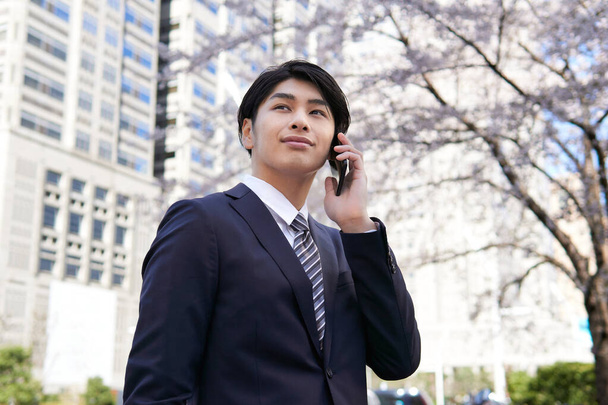 日本のビジネスマンがオフィス街の桜を背景にスマホで電話をかける. - 写真・画像
