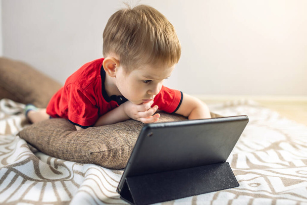 Een peuter kind kijkt tekenfilms op een tablet op het internet terwijl hij op een kussen op de vloer ligt. Gadgets en modern entertainment voor kinderen. - Foto, afbeelding