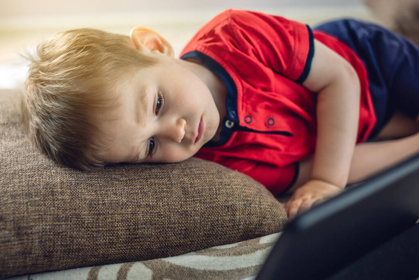A preschool toddler child watches cartoons on a tablet on the internet while lying on a pillow on the floor. Gadgets and modern entertainment for children. - Photo, image