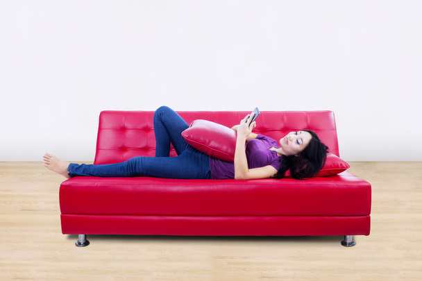 Attractive female on red sofa reading e-book at home - Photo, Image
