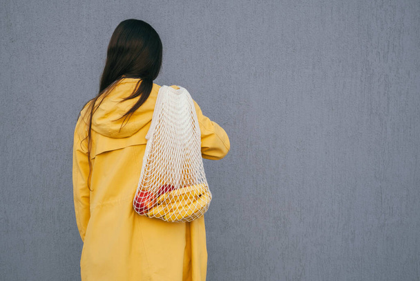 young woman with long dark hair wearing yellow raincoat holding string shopper bag with fruits standing back to the camera against gray wall - Photo, Image