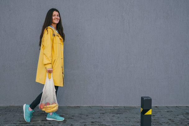 young woman in a yellow raincoat carrying string bag with fresh bananas and apples on the street - Foto, Imagen