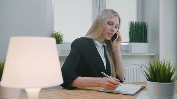 Ofiste akıllı telefondan konuşan bir iş kadını. Ofisteki zarif sarışın kadın iş yerinde oturuyor ve elinde cep telefonuyla gerekli bilgileri not defterine yazıyor. - Fotoğraf, Görsel
