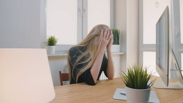 Mujer molesta sentada en el lugar de trabajo. Mujer rubia triste en ropa elegante agarrando la cabeza después del fracaso o malas noticias sentado en la oficina de luz en el escritorio con el ordenador
. - Foto, Imagen