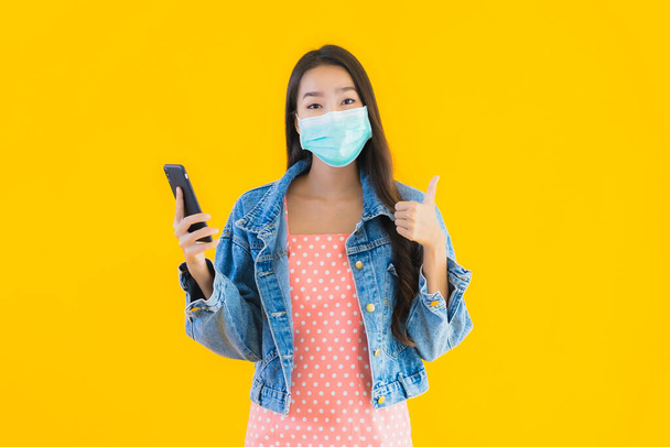 肖像画美しい若いアジアの女性は、保護するためのマスクを身に着けていますcovid19またはコロナウイルス黄色の隔離された背景にスマートフォンを使用 - 写真・画像