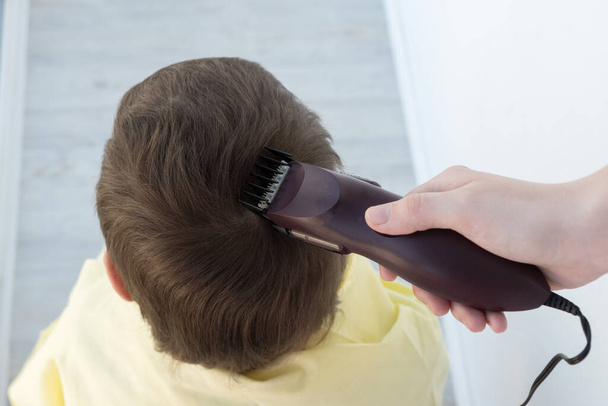 dłoń trzyma elektryczną maszynkę do strzyżenia włosów i obcina chłopcu długie włosy, fryzjer w domu, rodzic obcina włosy, podczas gdy fryzjerzy są zamknięci, zostaje w domu koncepcja - Zdjęcie, obraz