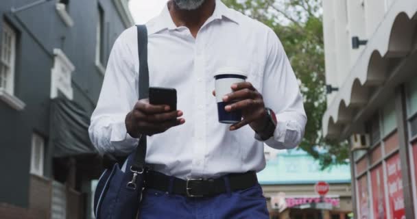 African American man out and about in the city streets during the day, walking down the street, drinking a takeaway coffee and using his smartphone. - Filmati, video