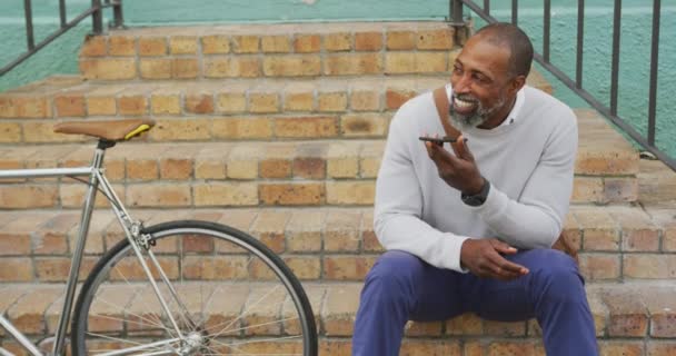 Uomo afroamericano in giro per le strade della città durante il giorno, seduto sulle scale, e parlare sul suo smartphone mentre un altro uomo è di passaggio e pugno urtando
. - Filmati, video
