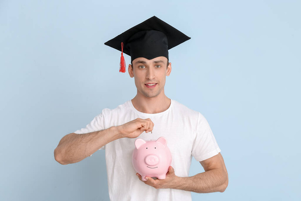 Young man in graduation hat and with piggy bank on light background. Tuition fees concept - Photo, Image