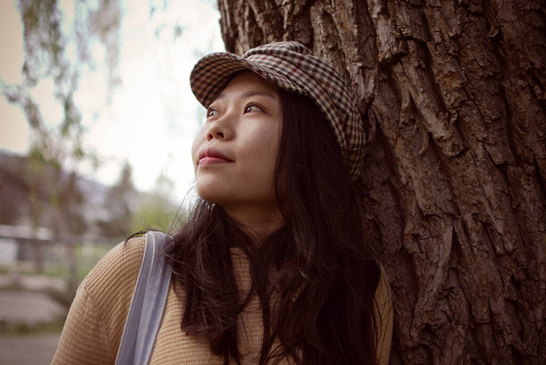Estudiante asiática universitaria internacional apoyada en un árbol mientras mira a la distancia pensativamente
 - Foto, imagen