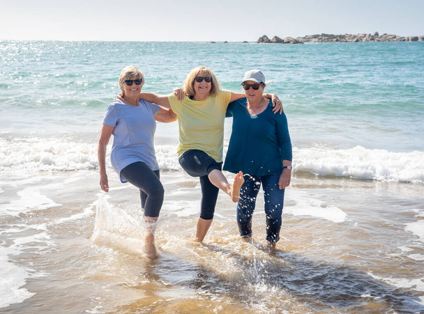 Lovely group of three senior retired woman on their 60s walking and having fun on the beach. Mature females Laughing and kicking water smiling playful enjoying retirement lifestyle and friendship. - Fotoğraf, Görsel