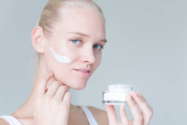 Modello femminile che idrata il viso tenendo un barattolo di crema di fronte a uno sfondo bianco con spazio copia. - Foto, immagini