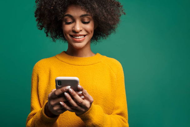 携帯電話を使って緑の背景に隔離されたセーターを着た若いアフリカ人女性の肖像画 - 写真・画像