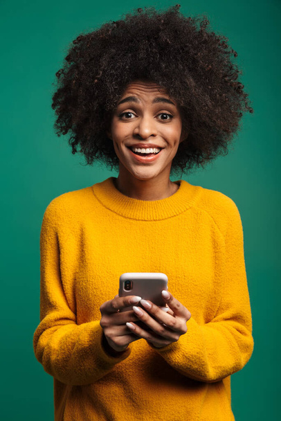 Portrait of a smiling young african woman wearing sweater standing isolated over green background, using mobile phone - Foto, Imagem