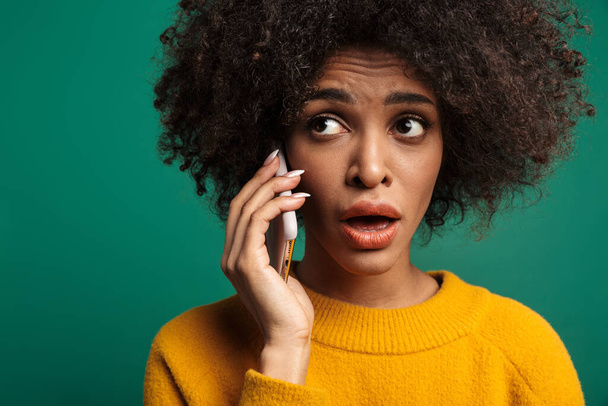 Portrait of a confused young african woman wearing sweater standing isolated over green background, talkning on mobile phone - Foto, afbeelding