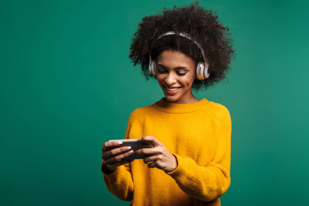Imagem de uma bela mulher africana encaracolado posando isolado sobre fundo de parede azul escuro ouvir música com fones de ouvido jogar jogos por smartphone
. - Foto, Imagem