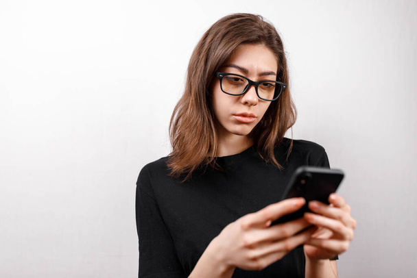 mooie jonge vrouw op een witte achtergrond kijkt naar de telefoon met bril en een zwart T-shirt. kopieerruimte. geïsoleerd - Foto, afbeelding