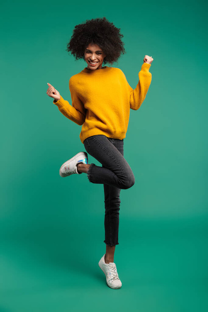 Full length portrait of a smiling young african woman wearing sweater jumping isolated over green background, celebrating success - Fotoğraf, Görsel