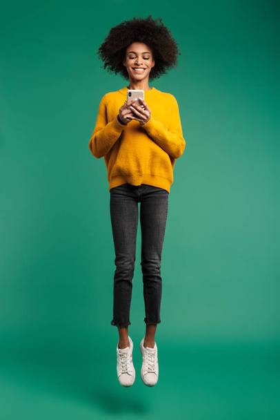 Full length portrait of a smiling young african woman wearing sweater jumping isolated over green background, using mobile phone - Foto, Imagem