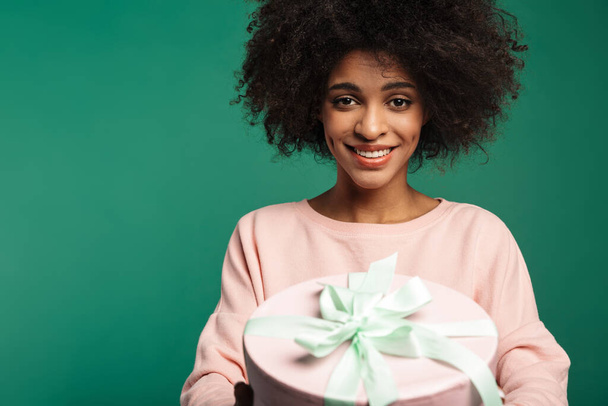 Porträt einer lächelnden jungen Afrikanerin in Sweatshirt, die isoliert vor grünem Hintergrund steht und eine große Geschenkschachtel in der Hand hält - Foto, Bild