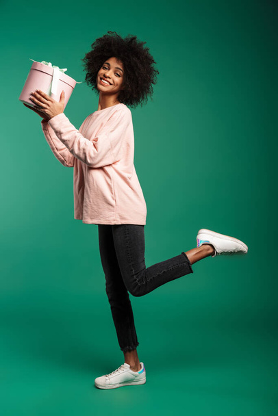 Full length portrait of a smiling young african woman wearing sweatshirt jumping isolated over green background, holding big present box - Foto, immagini